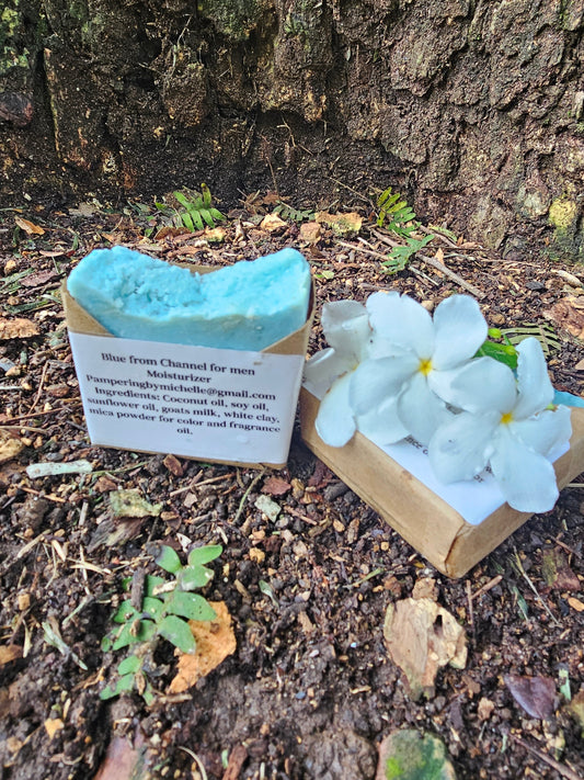 Blue men's soap bar in kraft packaging beside white flowers outdoors.
