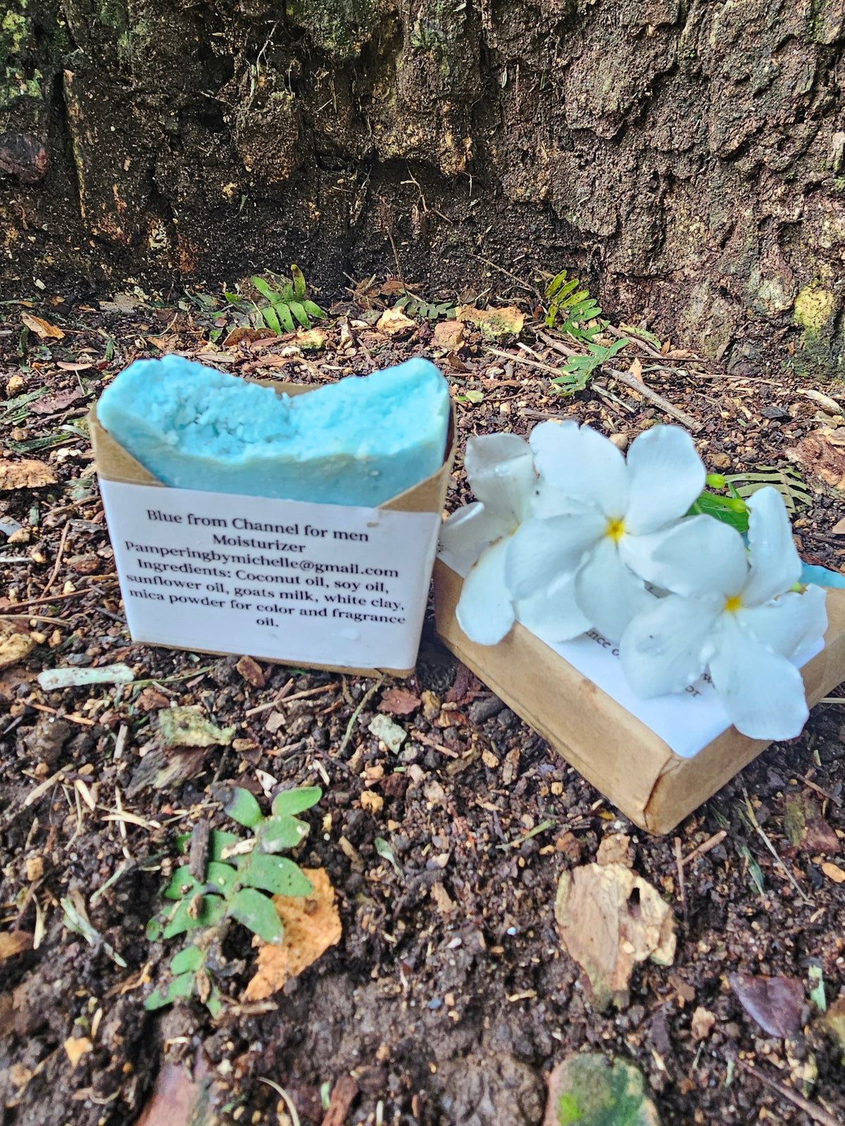 Blue men's soap bar in kraft packaging beside white flowers outdoors.